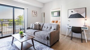 A modern living room with a grey sofa, a small table with a plant, a desk with a chair, and a large window with a view of the city. - Landing | Chic 1BD, Courtyard, Yoga Studio (Salt Lake City)