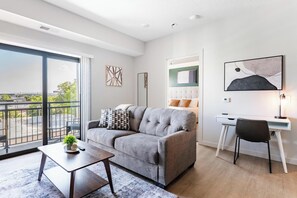 Ein modernes Wohnzimmer mit einem grauen Sofa, einem kleinen Tisch mit Pflanze, einem Schreibtisch mit Stuhl und einem großen Fenster mit Blick auf die Stadt.