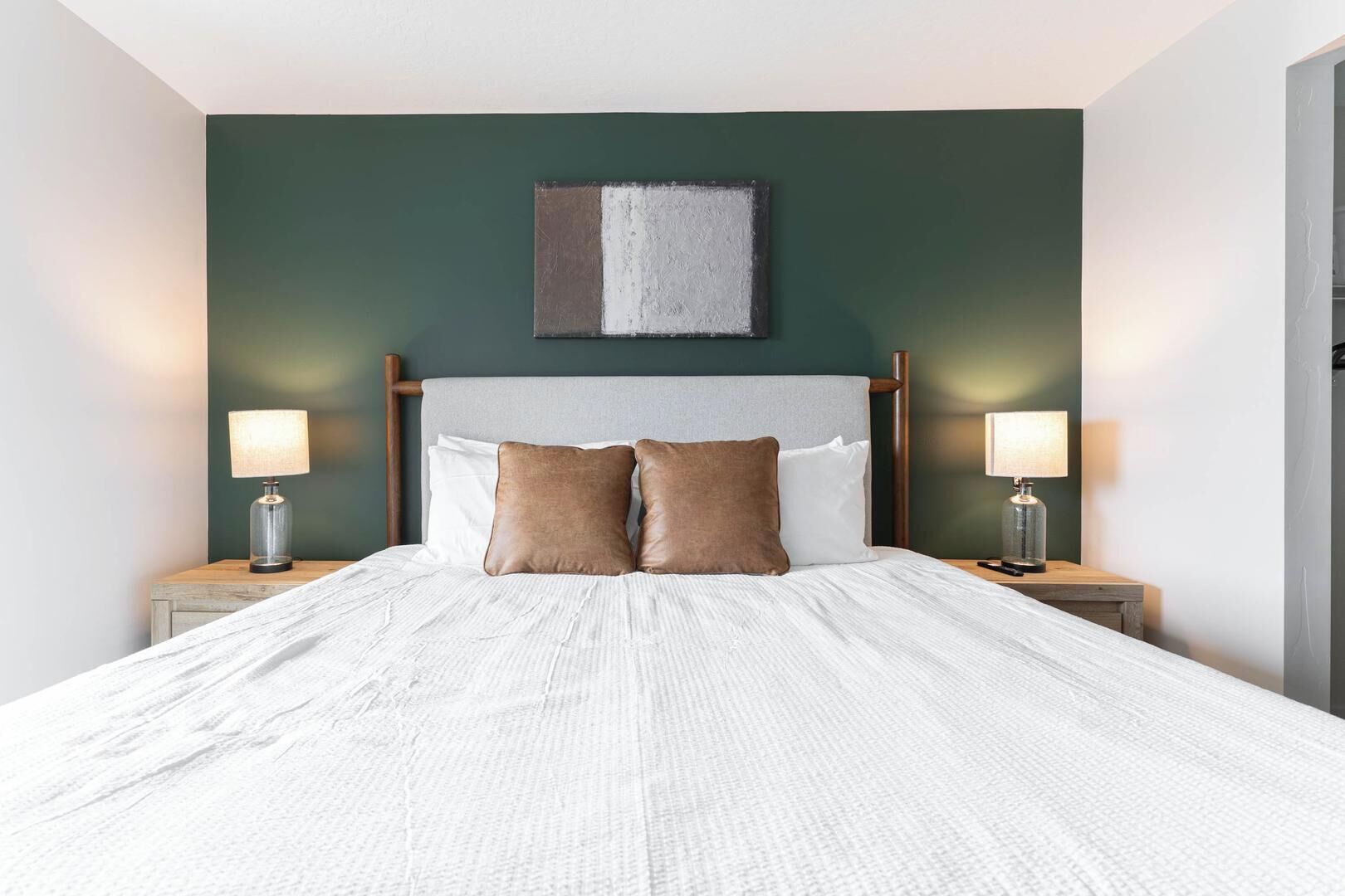 Een net opgemaakt bed met een witte deken en twee bruine kussens, met aan weerszijden nachtkastjes met lampen, in een kamer met een donkergroene wand en een eenvoudig, modern hoofdbord.