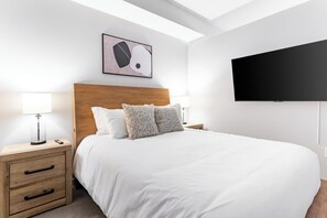 A hotel room with a bed, bedside tables, a lamp, a remote control, a wall-mounted TV, and a ceiling light fixture. - Landing | Stylish Studio, Yoga Studio, Gym (Salt Lake City)