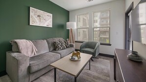 Ein Wohnzimmer mit einem grauen Sofa, einem kleinen Tisch und einem Sessel. Ein Bild hängt an der Wand, und neben dem Sofa steht eine Lampe.