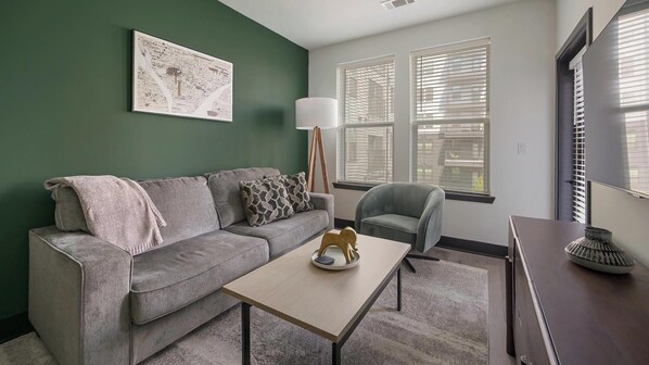 A living room with a grey sofa, a small table, and a chair. There is a painting on the wall and a lamp beside the sofa. - Landing | Stylish 1BD (Nashville)