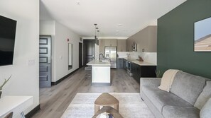 A modern kitchen with a central island, a dining area with a grey sofa, and a flat-screen TV mounted on the wall. - Landing | Spectacular 1BD (Nashville)