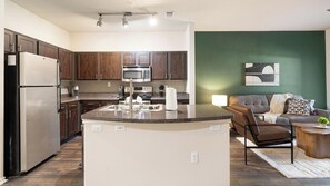 A modern kitchen with dark wood cabinets, stainless steel appliances, and a granite countertop island. - Landing | Stylish 1BD, Clubhouse (Irving)
