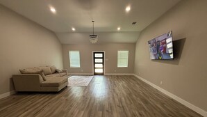 A spacious living room with a beige sofa, wooden flooring, and a large TV on the wall. - Matthews Blue house spacious 5BD and 2BA in calm neighborhood (Wilmer)