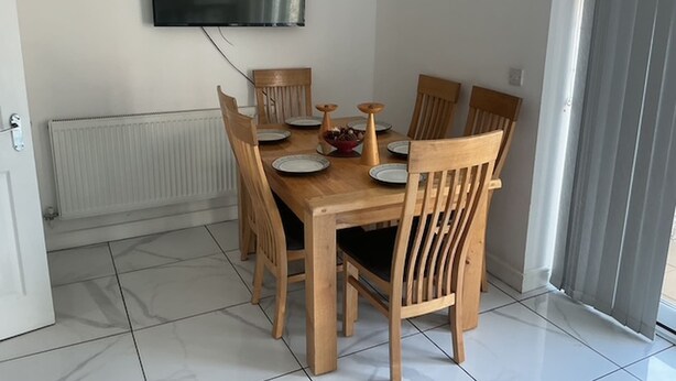 Un comedor con una mesa de madera, seis sillas, un televisor de pantalla plana en la pared y un radiador debajo.