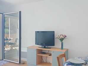 A room with a sliding glass door, a television on a wooden stand, a vase with flowers, and a dining table with plates and cutlery. - Coeur d'Oléron by Interhome (Ile d'Oléron)
