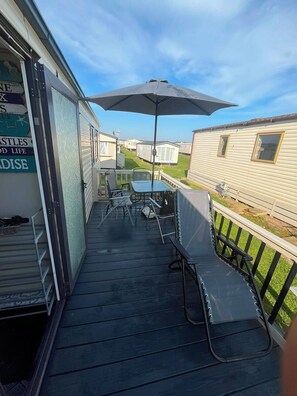 A covered outdoor area with a table and chairs, a large umbrella, and a view of adjacent mobile homes.
