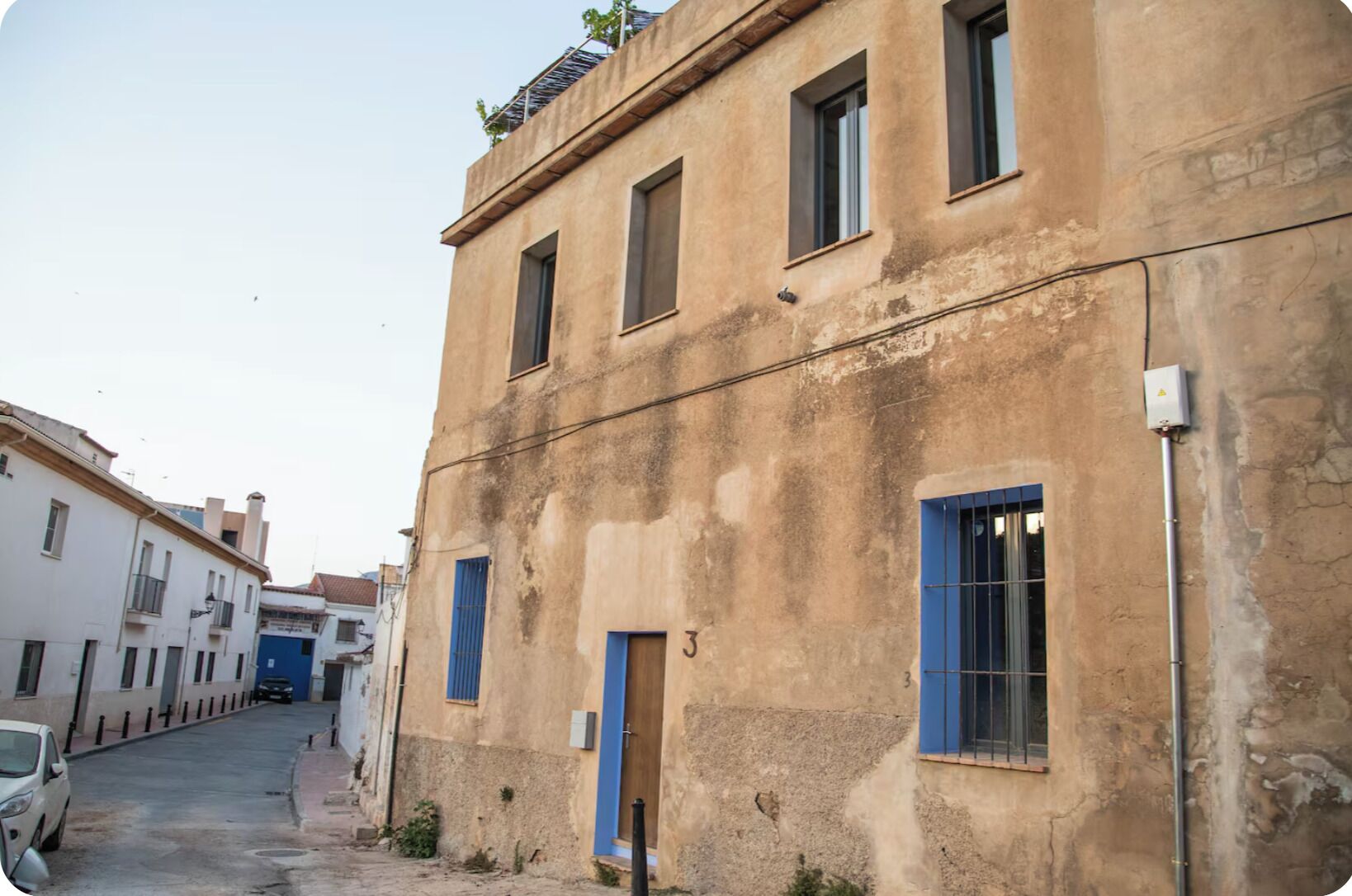 A narrow street with a building featuring a blue door and windows, adjacent to a white building with balconies.