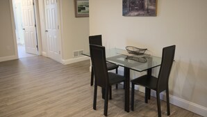 Uma sala de jantar com mesa de vidro e quatro cadeiras, um quadro na parede e uma porta para outro cômodo.