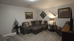 Uma sala de estar com um sofá seccional, uma mesinha com abajur, um espelho decorativo e um quadro na parede.