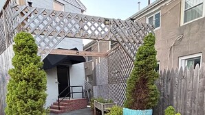 Um pátio com um arco de madeira treliçada, árvores coníferas em vasos e um edifício com fachada cinzenta.