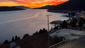 Vista costeira com pôr do sol sobre a água e montanhas, rua com calçada para pedestres e edifícios ao primeiro plano.