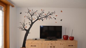 A room with a television on a wooden cabinet, a wall decal of a tree with leaves, and two red vases. - Alpenperle Appartement/Fewo 6-Bettwohnung by Interhome (Saas-Almagell)