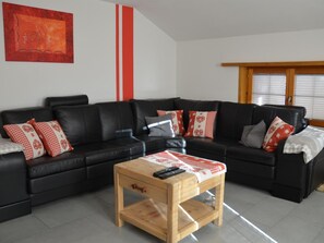 Een woonkamer met een zwarte leren hoekbank, een houten salontafel en kussens met een rood-wit patroon.