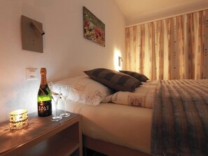 Une chambre à coucher avec un lit, des oreillers, une table de chevet avec une bouteille et des verres, un tableau au mur et des rideaux.