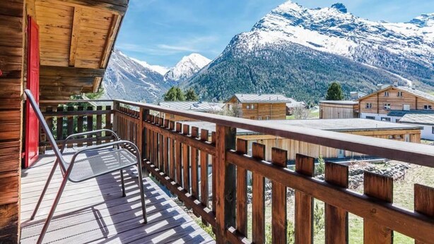 Ein Holzbalkon mit einem Metallstuhl, mit Blick auf einen Berg mit schneebedeckten Gipfeln und einem klaren Himmel.