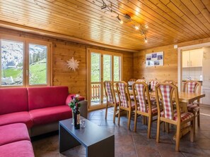 Een houten interieur met een rode bank, een eettafel en stoelen, grote ramen met uitzicht op groen, en een plafond met zichtbare balken en verlichting.
