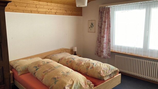 Huoneessa on puinen sänky, ikkunassa verhot ja seinässä patteri.