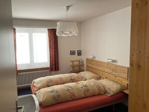 Habitación individual con una cama, cabecera de madera, ventana con cortinas, radiador y mesita de noche.