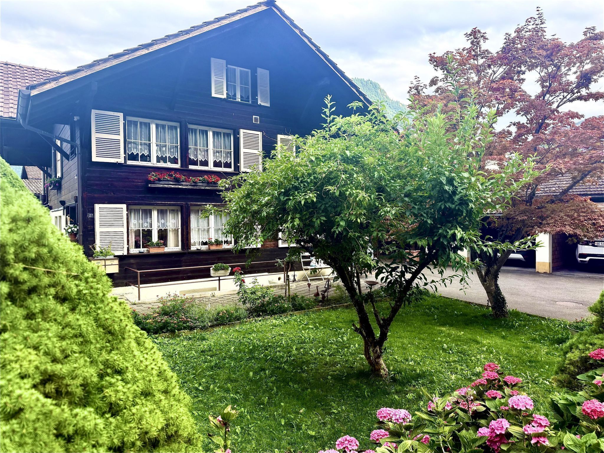 Ein zweistöckiges Gebäude mit dunkler Fassade, zahlreichen Fenstern mit Fensterläden und einem gepflegten Garten mit verschiedenen Pflanzen und Blumen.