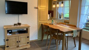 Un coin repas avec une table en bois, des chaises, un téléviseur fixé au mur et un espace cuisine visible à travers une porte ouverte.
