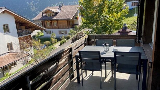 Un balcon avec une table et des chaises, donnant sur une région montagneuse parsemée de maisons.