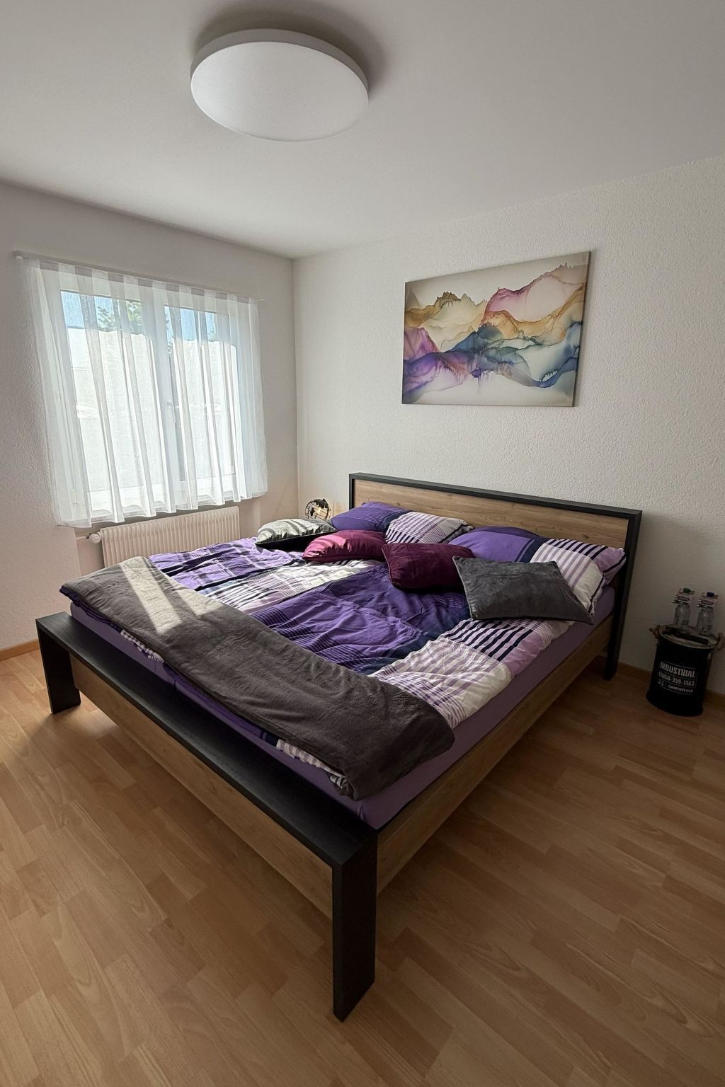 Ein Schlafzimmer mit Bett, einem Fenster mit transparenten Vorhängen, einem Heizkörper, einem Bild an der Wand und einem kleinen Tisch mit Flaschen.