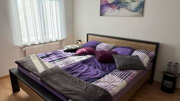 Ein Schlafzimmer mit Bett, einem Fenster mit transparenten Vorhängen, einem Heizkörper, einem Bild an der Wand und einem kleinen Tisch mit Flaschen.