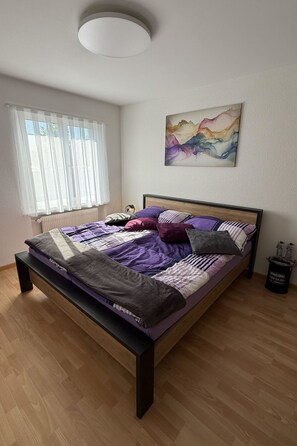 A bedroom with a bed, a window with sheer curtains, a radiator, a painting on the wall, and a small table with bottles.