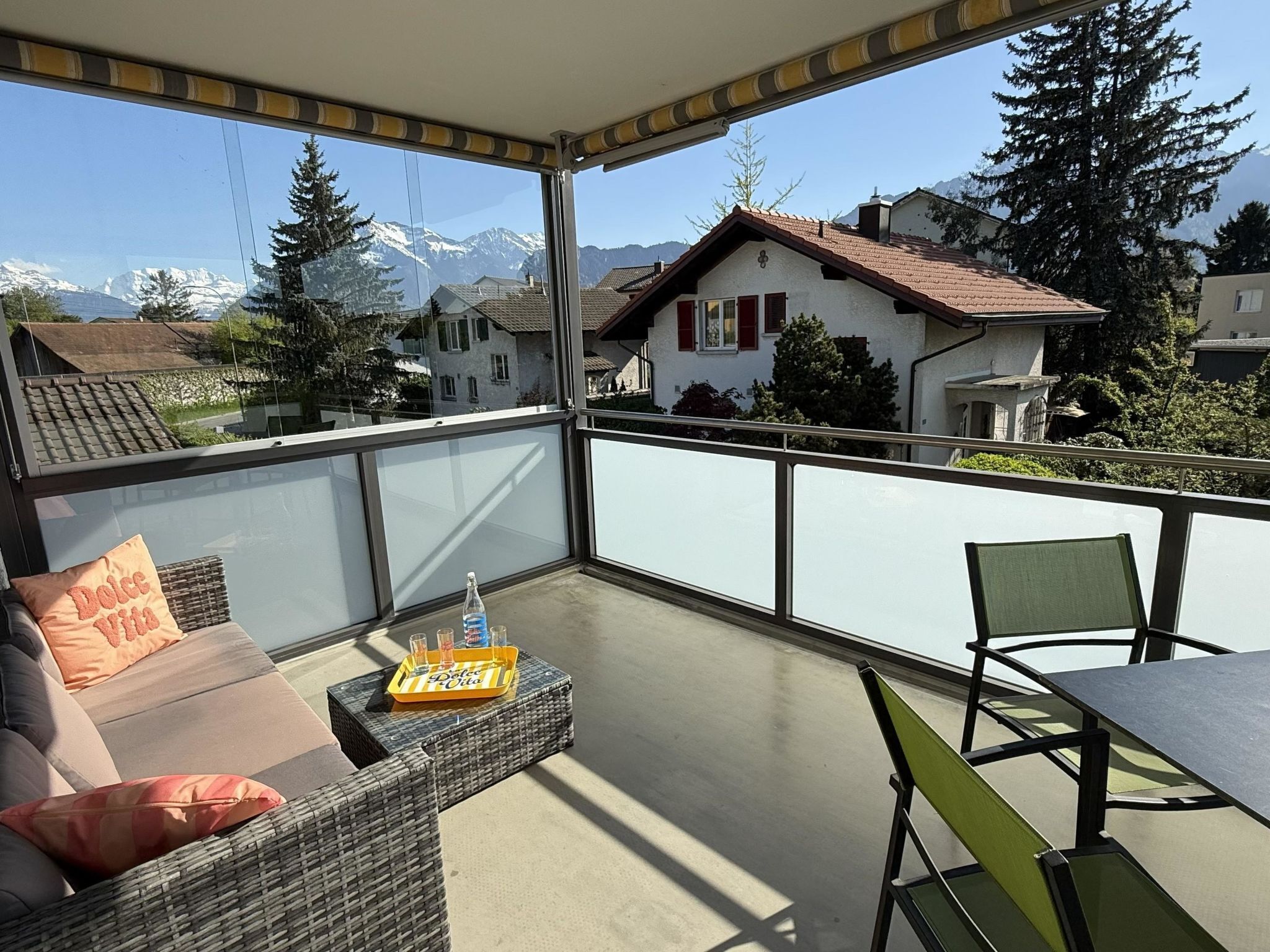 Ein Balkon mit einer Couch, einem Tisch und Stühlen, mit Blick auf ein Wohngebiet mit Häusern und Bergen.