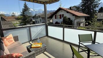 Ein Balkon mit einer Couch, einem Tisch und Stühlen, mit Blick auf ein Wohngebiet mit Häusern und Bergen.