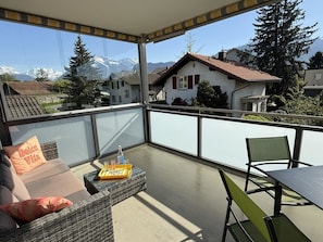 A balcony with a sofa, a table, and chairs, overlooking a residential area with houses and mountains.