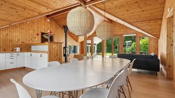 Ein geräumiges Zimmer mit einem großen runden Tisch, weißen Stühlen, einem schwarzen Sofa und einer Holzzieldecke mit Pendelleuchten.