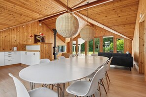A spacious room with a large round table, white chairs, a black sofa, and a wooden ceiling with hanging light fixtures.