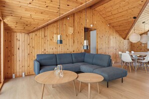 A modern interior with a wooden ceiling, a sectional sofa, two round tables, and a dining area with chairs.