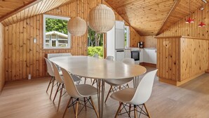 Een houten interieur met een grote eettafel, witte stoelen, hangende rieten lampen en een keukenhoek.