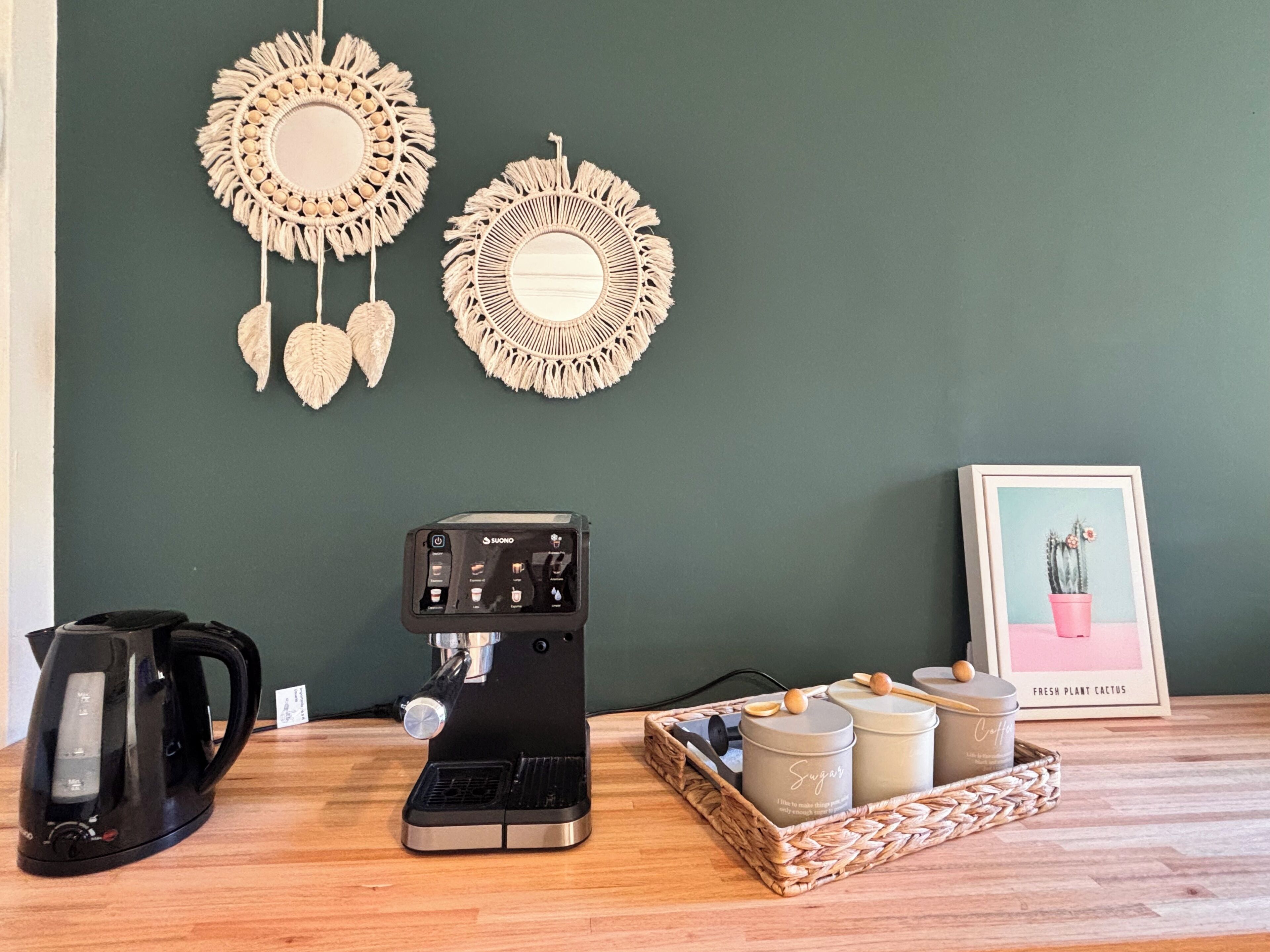 Una máquina de café, un hervidor y una bandeja con recipientes de azúcar y crema sobre una mesa de madera frente a una pared verde.