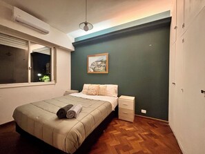 A bedroom with a bed, a nightstand, a dresser, a window, and an air conditioning unit. - Meraki: Bright & Cozy | Walk Everywhere (Buenos Aires)