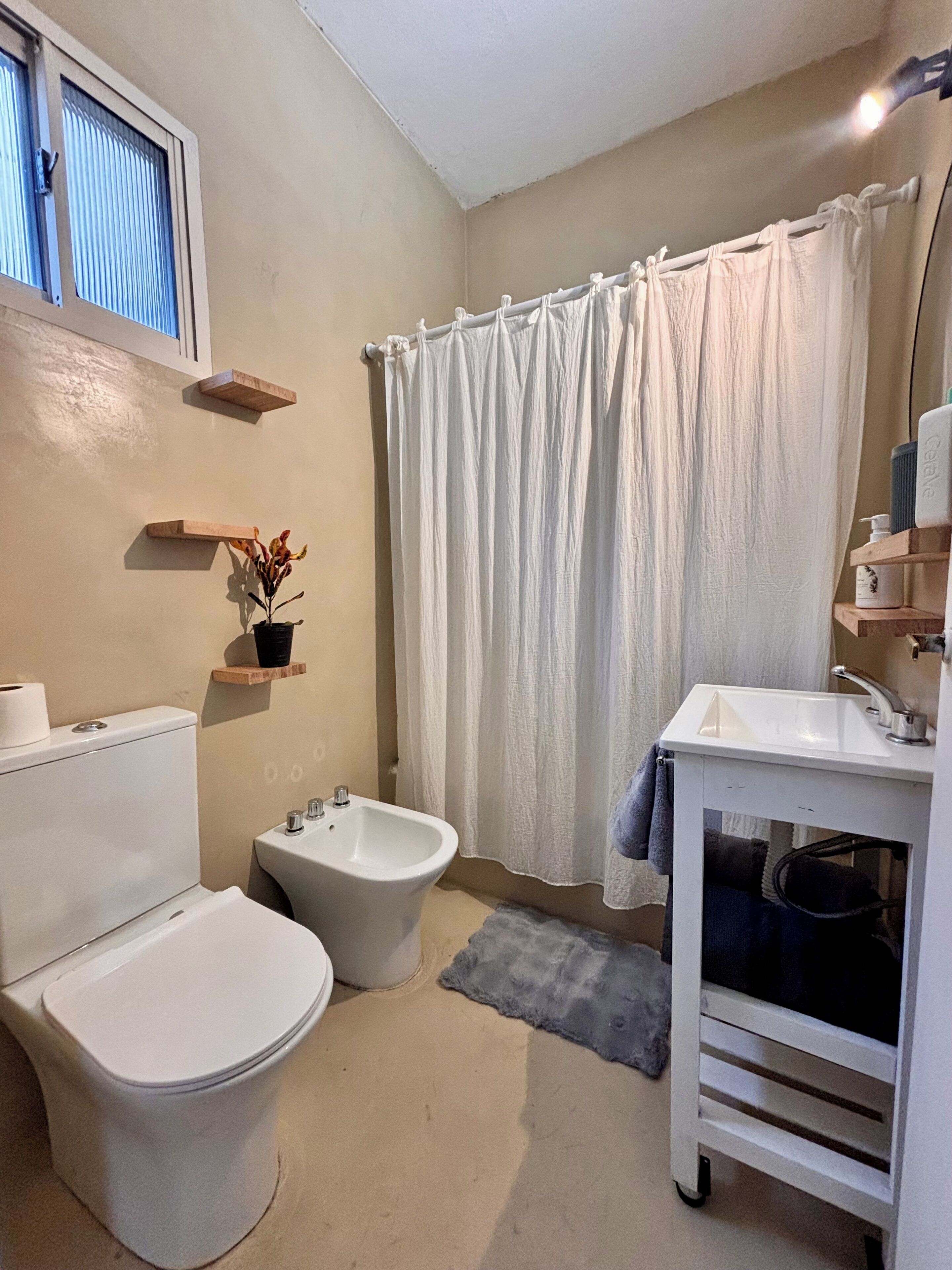 Un baño con inodoro, bidé y lavamanos. Se ven estantes de madera con una planta y artículos de aseo.