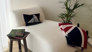 Ein gemütliches Zimmer mit einem weißen Sofa, einem kleinen Holzschemel mit einem Buch, einer Topfpflanze und einer Decke mit einem Flaggenmuster.