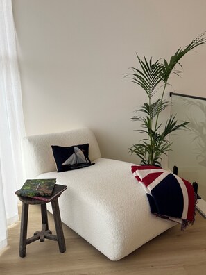 Ein gemütliches Zimmer mit einem weißen Sofa, einem kleinen Holzschemel mit einem Buch, einer Topfpflanze und einer Decke mit einem Flaggenmuster.