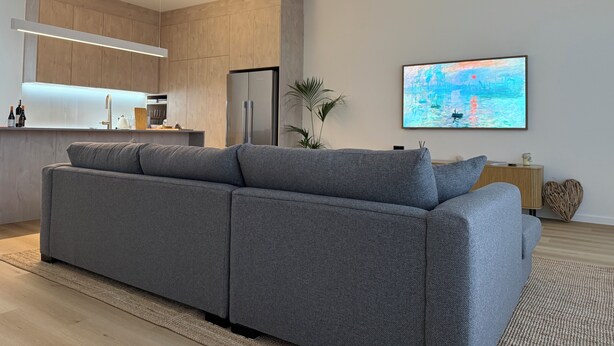 Ein modernes Wohnzimmer mit einem grauen Ecksofa, einem Flachbildfernseher und einer Küchenzeile mit Holzausbau und einem Edelstahlkühlschrank.
