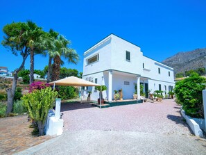Ein weißes Gebäude mit überdachter Terrasse, Palmen und einer hügeligen, felsigen Landschaft im Hintergrund.