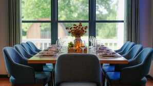 Een eettafel gedekt voor zes personen, met blauwe stoelen, een bloevas en uitzicht op bomen door grote ramen.