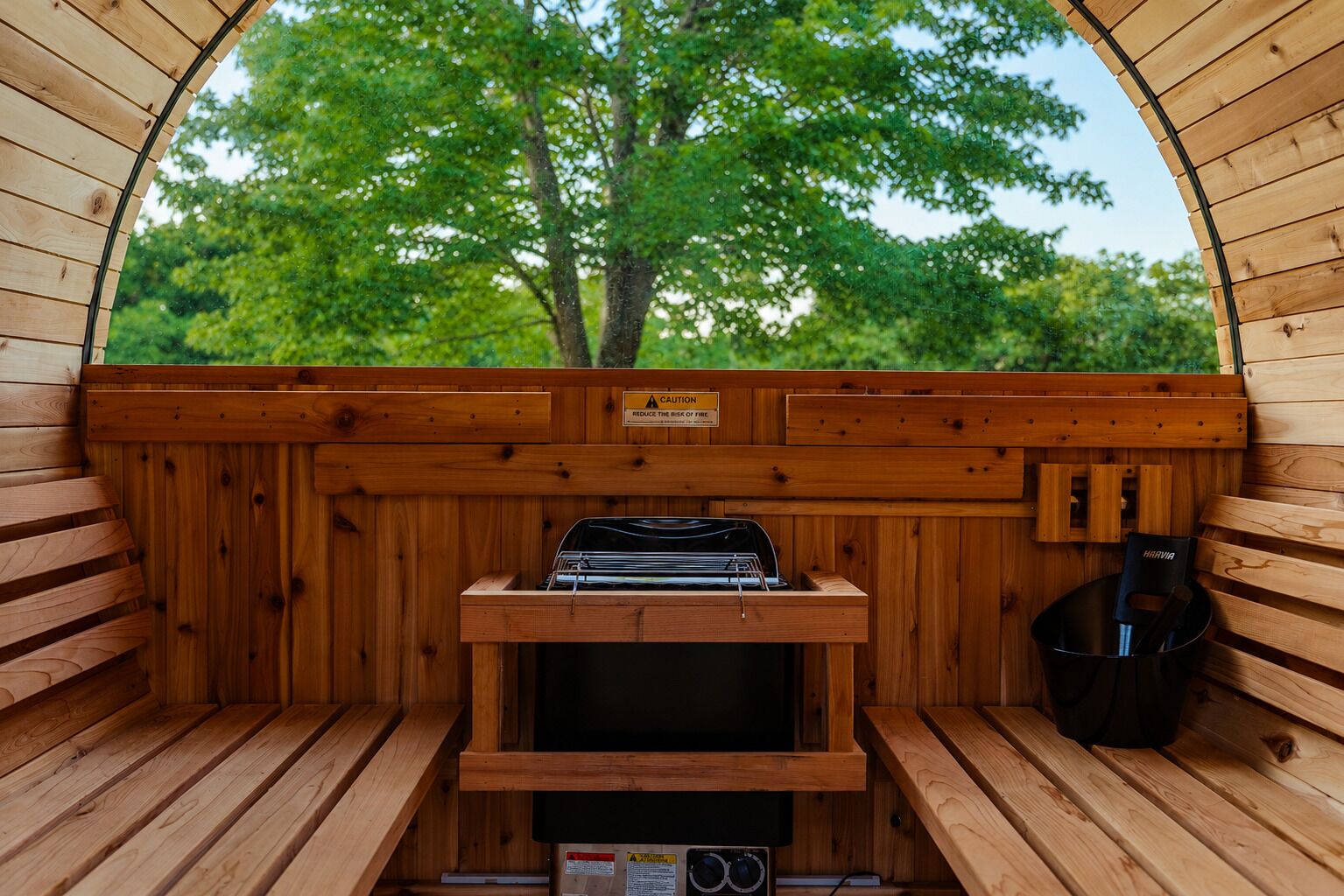 A sauna with wooden walls, a bench, a table with a grill, and a view of trees.