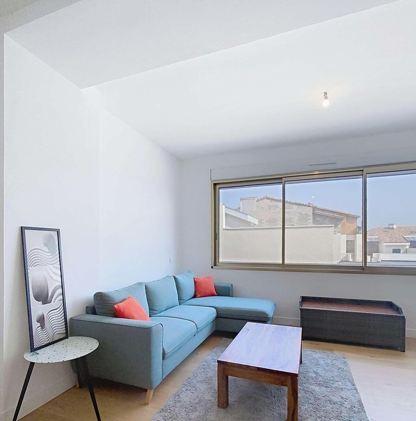 Ein modernes Wohnzimmer mit einem blauen Sofa, einem kleinen runden Tisch, einem Holztisch und einem großen Fenster mit Blick auf Gebäude.