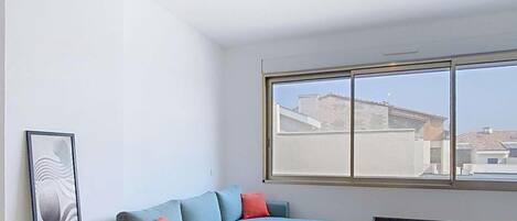 Ein modernes Wohnzimmer mit einem blauen Sofa, einem kleinen runden Tisch, einem Holztisch und einem großen Fenster mit Blick auf Gebäude.