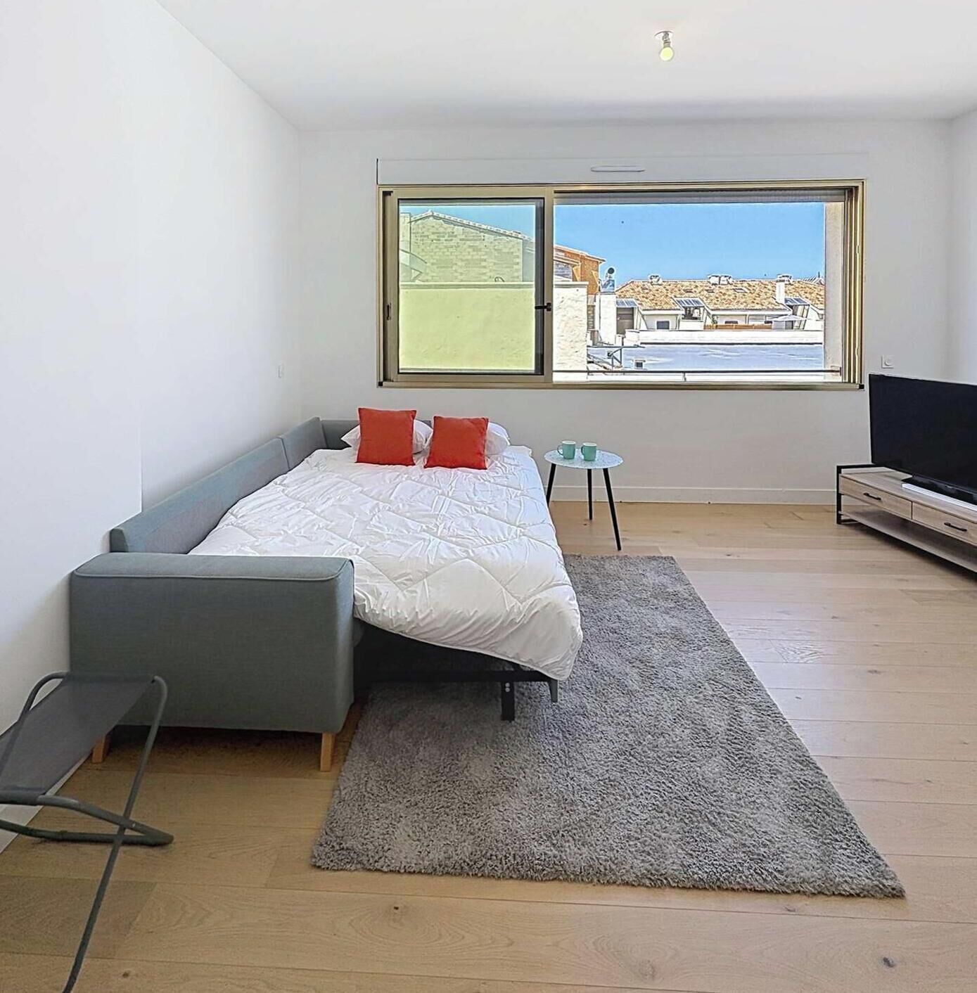 A modern bedroom with a bed, a grey sofa, a small table with cups, a TV, and a large window with a view of buildings and a clear sky.