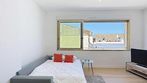 A modern bedroom with a bed, a grey sofa, a small table with cups, a TV, and a large window with a view of buildings and a clear sky. - Holiday studio close to the ocean – Ideal for 2 people (LACANAU OCEAN)